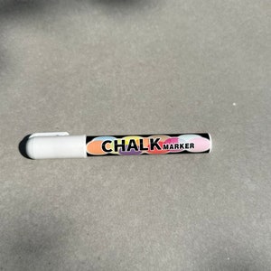 Può includere: Un pennarello a gesso bianco con un corpo nero. Il corpo presenta la scritta "CHALK MARKER" in un design colorato. Il pennarello è appoggiato su una superficie grigia.