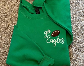 May include: A bright green crewneck sweatshirt featuring white embroidered cursive text that reads go Eagles, accompanied by an embroidered brown and white football graphic positioned on the upper right chest area of the garment.