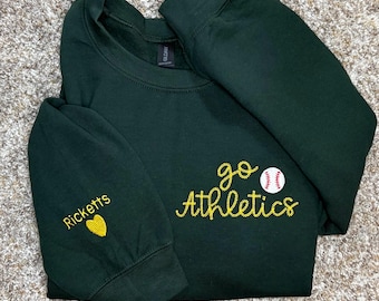 May include: A dark green long-sleeve sweatshirt featuring yellow embroidered text that reads go Athletics with a white baseball graphic. The left sleeve is embroidered with the name Ricketts in yellow text above a yellow heart icon.