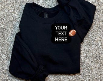 May include: A black long-sleeve sweatshirt featuring a rounded neckline and a black rectangular patch on the chest. The patch displays the white text "YOUR TEXT HERE" accompanied by a small graphic of a brown and white American football.