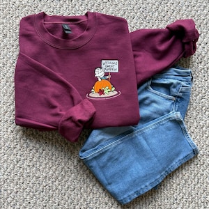 May include: A maroon crewneck sweatshirt featuring an embroidered graphic of a cartoon character sitting on a large orange pumpkin and holding a sign that reads "WELCOME GREAT PUMPKIN," paired with a pair of blue denim jeans.