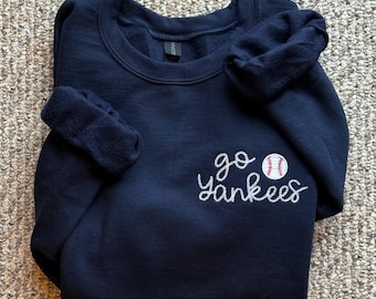 Op de afbeelding: Navyblauwe trui met witte geborduurde tekst die "go yankees" zegt met een rode en witte baseball afbeelding.