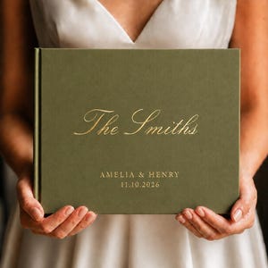 May include: A rectangular olive green photo album with a hard cover. The front cover features gold foil lettering that reads The Smiths, followed by the names AMELIA & HENRY and the date 11.10.2026 centered in a clean, sans-serif font.