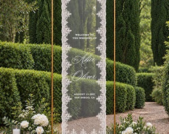 May include: A tall, rectangular, sheer white lace wedding sign hanging from a gold metal frame. The sign features the text: Welcome to the wedding of Alice & Oliver, August 15, 2027, San Diego, CA, surrounded by an intricate lace border.