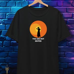 Puede incluir: Camiseta negra con un gráfico de una silueta contra un atardecer naranja y amarillo. El texto "YOU ARE NOT INVITED" está debajo. La camiseta está colgada en una percha de madera.