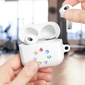 May include: White wireless earbuds and charging case. The case features colorful circular graphics. One earbud is held separately. The case is held in a person's hand. The background is blurred.