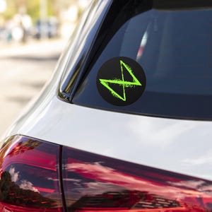 May include: A green and black sticker with a stylized lightning bolt design. The sticker is on the back window of a white car.