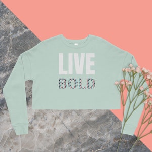 Könnte beinhalten: Ein hellgrüner, kurzer Pullover mit langen Ärmeln. Der Pullover hat den Schriftzug "LIVE BOLD" in Weiß auf der Vorderseite und ein geometrisches Muster in Rosa, Blau und Grau.
