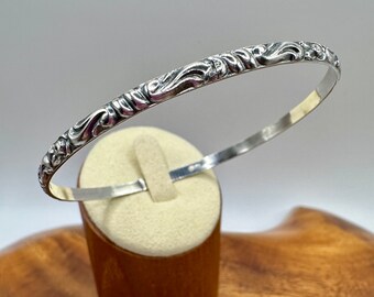 May include: A silver bangle bracelet with an intricate floral design. The bracelet is displayed on a wooden stand.