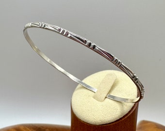 May include: A silver bangle bracelet with a geometric design. The bracelet is displayed on a small, cream-colored stand. The bangle has a series of rectangular and linear shapes. The background is a light gray.