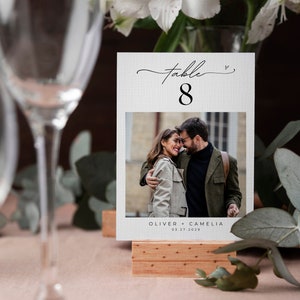 May include: White table number card with black text and a photo of a couple. The card reads "Table 8" and "Oliver + Camelia 03.27.2023".