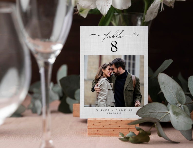 May include: White table number card with black text and a photo of a couple. The card reads "Table 8" and "Oliver + Camelia 03.27.2023".