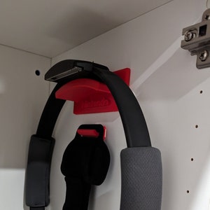 Peut inclure: Un anneau de Pilates noir avec une sangle rouge et noire suspendu à un mur blanc. L'anneau est fixé à un crochet en plastique rouge avec le mot "Nintendo" imprimé dessus.