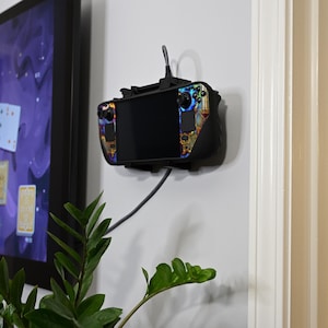 May include: A black wall-mounted gaming console holder with a gaming device attached. The device has a black screen and colourful accents. A cable extends from the device. A television displaying a card game is in the background.