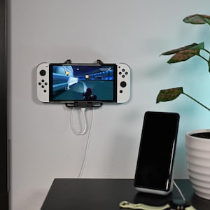 Op de afbeelding: Een witte Nintendo Switch-console is aan een muur bevestigd met een zwarte oplaadkabel. De console speelt een racespel. Een zwarte smartphone staat op een oplaadstandaard op een zwarte tafel. Een witte potplant staat op de tafel.