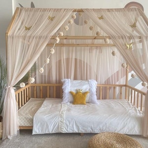 May include: A wooden bed frame with a canopy made of sheer fabric. The canopy is decorated with pom-poms and butterflies. The bed is made up with white bedding and a yellow pillow with a crown.