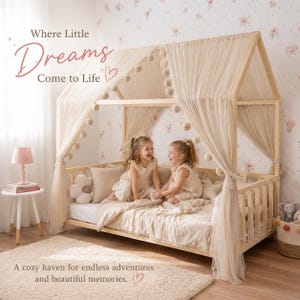 May include: A wooden canopy bed with sheer fabric and pom-pom accents. Two young girls in matching dresses sit on the bed, laughing. The room has floral wallpaper and a small side table with a lamp. Text reads: "Where Little Dreams Come to Life."