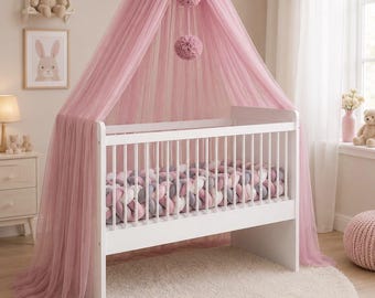 May include: A white crib with a pink tulle canopy and pom-pom accents. The crib is set on a cream-colored, round rug. A framed bunny print and a teddy bear are on the wall, creating a soft, nursery theme.