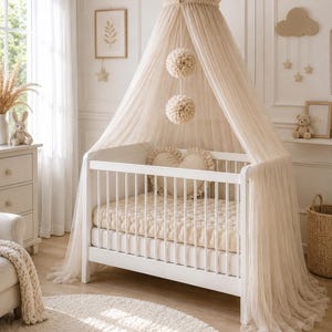Könnte beinhalten: Ein weißes Babybett mit einer cremefarbenen Matratze und einem transparenten Baldachin. Zwei dekorative Pompons hängen am Baldachin. Der Raum ist in neutralen Tönen gehalten, mit einem runden Teppich und einem Bilderrahmen.