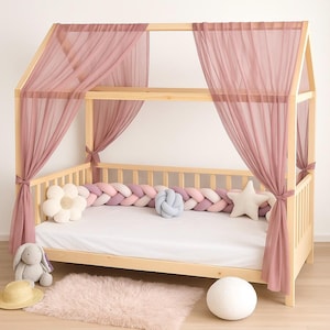 Könnte beinhalten: Ein hölzernes Himmelbett mit transparenten, staubrosa Vorhängen. Das Bett ist mit einem geflochtenen Stoßfänger in Rosa- und Weißtönen und dekorativen Kissen ausgestattet. Ein Plüschteppich, ein Stoffhase und andere dekorative Gegenstände vervollständigen die Szene.