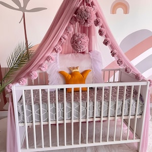 May include: A white crib with a pink tulle canopy and pom poms. The canopy is decorated with gold butterflies. A white pillow with a gold crown is on the crib mattress.