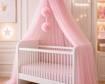 May include: A white crib with a pink canopy and bedding. The canopy is made of sheer pink fabric and adorned with two pink pom-poms. A pink rug with a crown design is on the floor. The room is decorated in soft pink tones, creating a nursery theme.