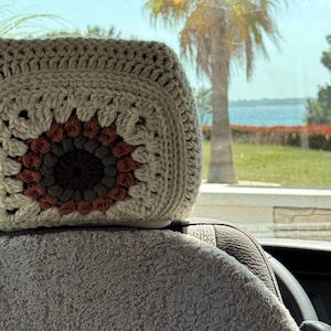May include: A crocheted headrest cover in a car. The cover features a square granny square design with a cream-coloured border and a central pattern of brown, green, and cream. The headrest is on a car seat with a grey fuzzy cover.
