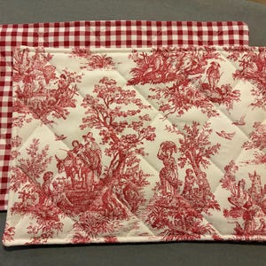 May include: Two quilted placemats. One features a red and white gingham pattern. The other has a red toile design on a white background, depicting pastoral scenes. Both are rectangular.