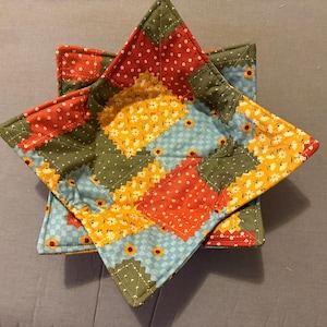 Può includere: Un sottociotola quadrato in tessuto con un design patchwork caratterizzato da quadrati alternati rossi a pois bianchi, gialli con piccoli girasoli, verde oliva a pois bianchi e azzurri a quadretti, il tutto tenuto insieme da cuciture bianche.