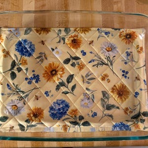May include: A rectangular clear glass baking dish containing a quilted fabric hot pad. The hot pad features a light yellow background with a floral pattern of blue hydrangeas, white daisies, and yellow sunflowers, accented by small bees.