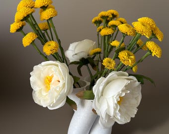 May include: White ceramic vase with a unique, organic shape, holding a bouquet of yellow button flowers and white peonies.