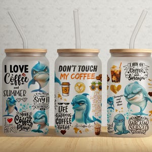 Puede incluir: Tres vasos de vidrio transparente con tapa de madera y una pajita. Cada vaso tiene un diseño diferente con un delfín y texto. El primer vaso dice "Me encanta el café y el verano". El segundo vaso dice "No toques mi café". El tercer vaso dice "Funciono con café y sarcasmo".