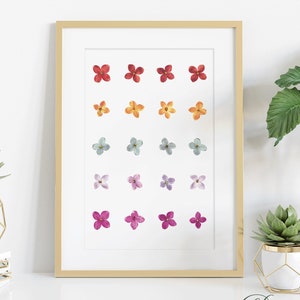 以下が含まれることがあります： 水彩の花の虹色のグラデーションが描かれた額装されたプリント。プリントは白い棚に置かれ、2つの鉢植えの植物が置かれています。