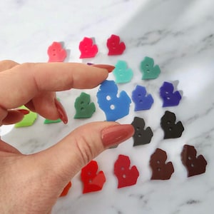 May include: A collection of colorful Michigan-shaped buttons in various shades, including red, blue, green, and brown. The buttons are small and have two holes. A hand is holding a blue button.