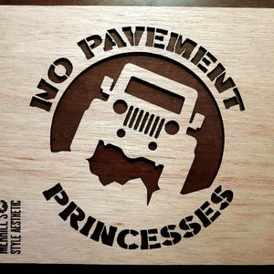 May include: Wooden sign with a Jeep silhouette inside a brown circle, surrounded by the words "NO PAVEMENT PRINCESSES." The sign also includes the text "MERRILL'S STYLE AESTHETIC" in the lower left corner. The sign is made of light-colored wood.