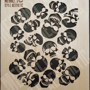 May include: A rectangular light-colored wooden stencil sheet featuring multiple cut-out skull designs of varying sizes and orientations. The top left corner displays the text MERRILL'S STYLE AESTHETIC. The skulls have detailed teeth and eye sockets.