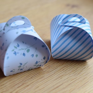 以下が含まれることがあります： 2つの小さな装飾用の紙製ギフトボックス。1つは青い花の小枝が付いた白で、もう1つは青と白の斜めストライプです。どちらも上部に小さなハンドルがあり、木製の表面にあります。