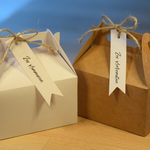 Könnte beinhalten: Zwei Geschenkboxen, eine weiße und eine braune, sind mit Kordelbändern gebunden. Jede Box hat ein Etikett mit deutschem Text. Die weiße Box sagt "Zur Kommunion" und die braune Box sagt "Zur Konfirmation".