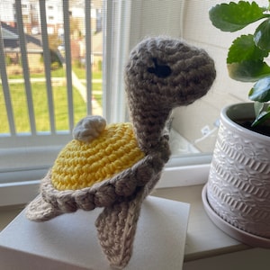 May include: A crocheted turtle toy with a yellow shell and a brown head and legs. The turtle is sitting on a white box.