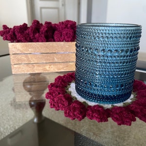 May include: A blue glass with a textured surface sits on a crocheted coaster with a burgundy flower design.