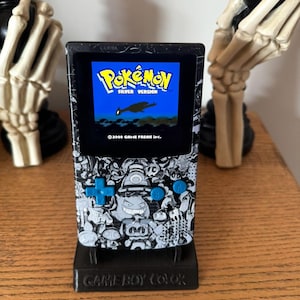 May include: A black and white Game Boy Color with a custom Pokemon Silver Version skin. The screen shows the game's title screen. The Game Boy Color is on a black stand with the words "Game Boy Color" printed on it.