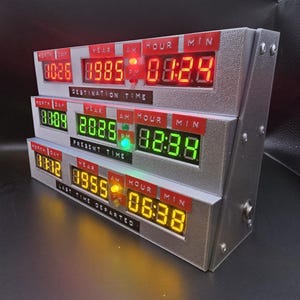 May include: A silver-colored electronic time display unit featuring three horizontal rows of digital LED readouts. The top row displays red numbers labeled Destination Time, the middle row shows green numbers for Present Time, and the bottom row displays yellow numbers for Last Time Departed, each with red labels for Month, Day, Year, Hour, and Minute.