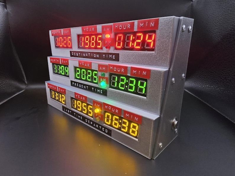 May include: A silver-colored electronic time display unit featuring three horizontal rows of digital LED readouts. The top row displays red numbers labeled Destination Time, the middle row shows green numbers for Present Time, and the bottom row displays yellow numbers for Last Time Departed, each with red labels for Month, Day, Year, Hour, and Minute.