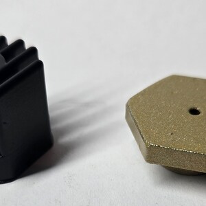 Op de afbeelding: Twee kleine zwarte en gouden metalen voorwerpen. Het zwarte voorwerp heeft een geribbeld ontwerp en het gouden voorwerp is zeshoekig met een klein gaatje in het midden.