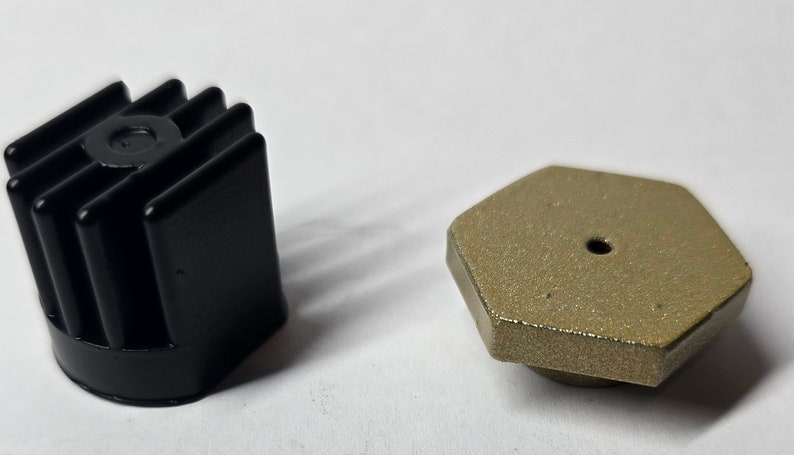 Op de afbeelding: Twee kleine zwarte en gouden metalen voorwerpen. Het zwarte voorwerp heeft een geribbeld ontwerp en het gouden voorwerp is zeshoekig met een klein gaatje in het midden.