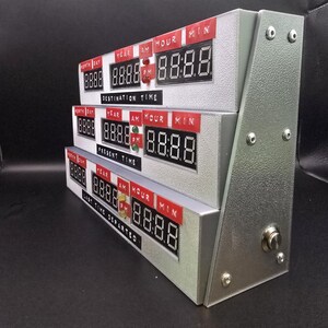 May include: A silver-colored time display unit featuring three stacked digital clock modules. Each module has red labels for Month, Day, Year, AM, Hour, and Min, with black digital screens. The labels read Destination Time, Present Time, and Last Time Departed.