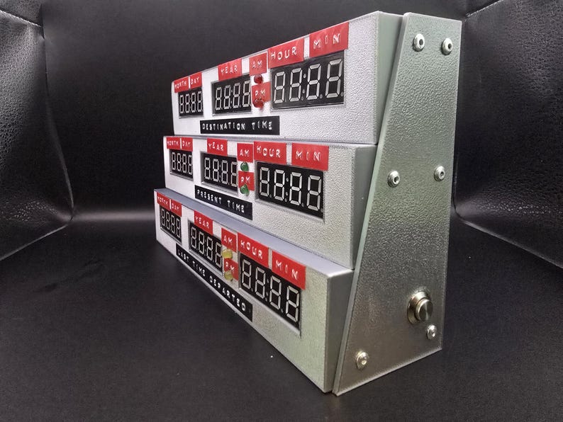 May include: A silver-colored time display unit featuring three stacked digital clock modules. Each module has red labels for Month, Day, Year, AM, Hour, and Min, with black digital screens. The labels read Destination Time, Present Time, and Last Time Departed.