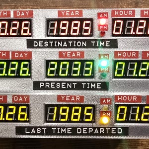 May include: A silver-toned time display unit featuring three horizontal rows of digital LED readouts labeled Destination Time, Present Time, and Last Time Departed. Each row displays Month, Day, Year, Hour, and Minute with red, green, and yellow lights.