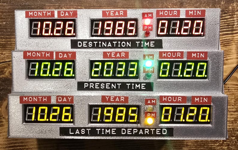 May include: A silver-toned time display unit featuring three horizontal rows of digital LED readouts labeled Destination Time, Present Time, and Last Time Departed. Each row displays Month, Day, Year, Hour, and Minute with red, green, and yellow lights.