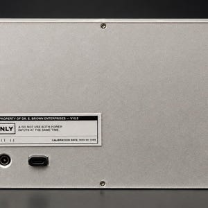 May include: A rectangular silver metal electronic device enclosure with four corner screws. A white label reads: PROPERTY OF DR. E. BROWN ENTERPRISES &mdash; V10.5, 5V ONLY, A DO NOT USE BOTH POWER INPUTS AT THE SAME TIME, CALIBRATION DATE: NOV 05 1955. Two power ports are visible.